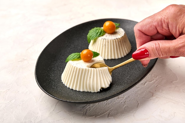 Woman39s hand with spoon breaks off piece of homemade panna cotta with natural vanilla pod physalis or winter cherry and mint on black plate