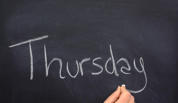 Woman39s hand writing the day of the week on a blackboard with white chalk Thursday