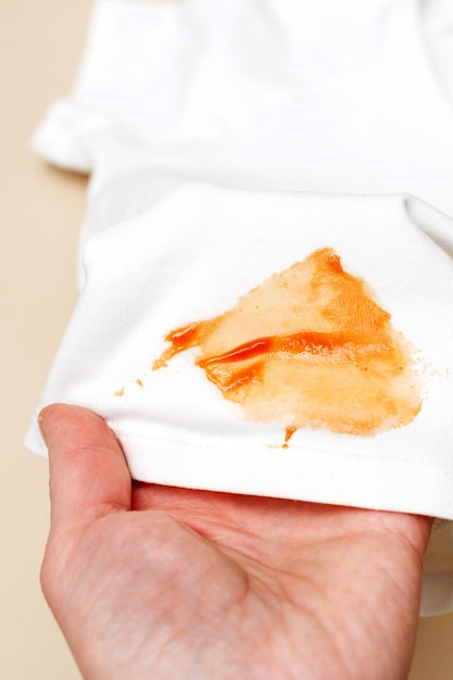 Womans hand hold white shirt clothes with ketchup stain on a beige background