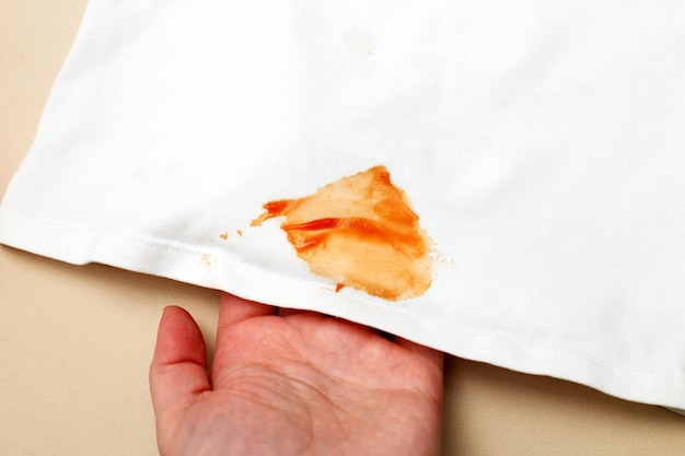 Womans hand hold white shirt clothes with ketchup stain on a beige background