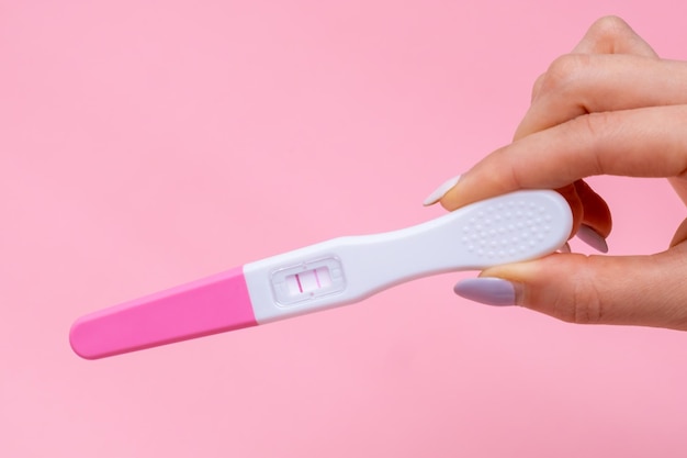 Womans hand holding a positive pregnancy test with two stripes on pink background with copy space