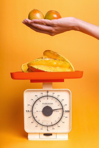 Womans hand holding tomatoes with starfruits on weigh scales over yellow background