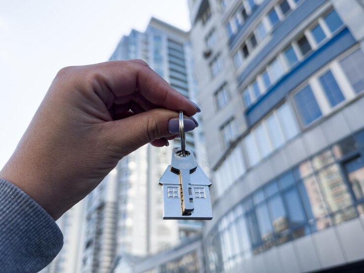 A womans hand holds a house key in plain view in front of a tall elegant modern building rising