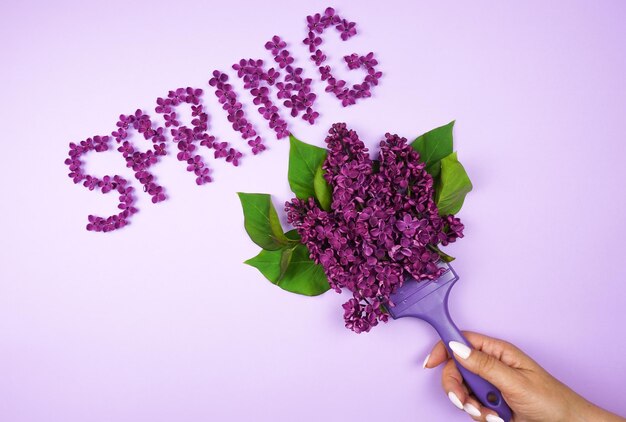 A womans hand holds a purple paint brush and paints the word spring with lilac flowers