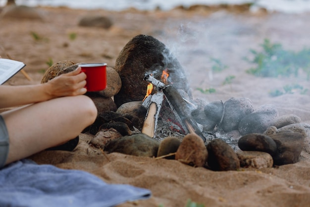 A womans hand holds a red cup of coffee or tea against the background of a campfire on a hike