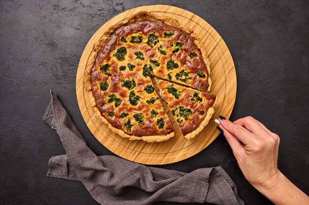 Womans hand holds spatula with piece of quiche tart with broccoli and tuna on round wooden cutting