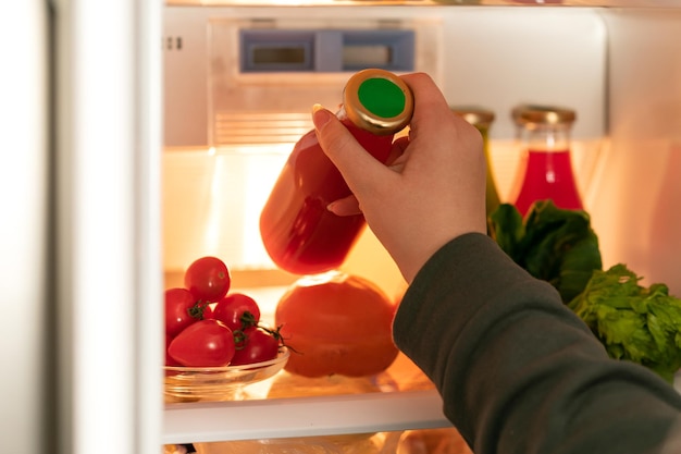 a womans hand pulls out a glass bottle with red juice from the refrigerator