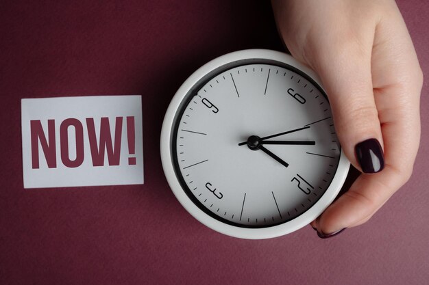 Womans hand show an alarm clock and a paper with the inscription NOW 
Now is written on a paper