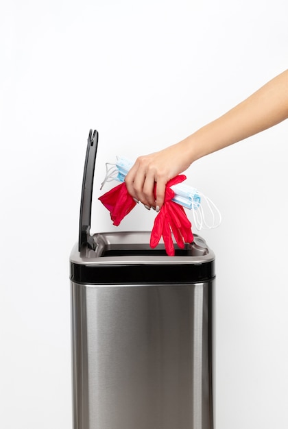 Womans hand throws a protective mask and rubber gloves into the trash can on a gray background