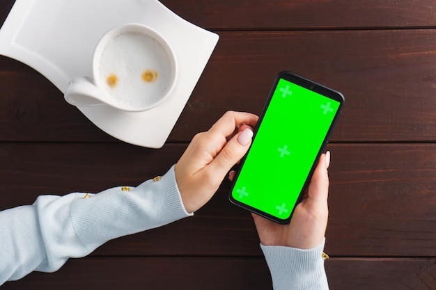 女性の手はコーヒーカップとカフェのテーブルでオンラインショッピングのために緑色の画面のスマートフォンを使用します