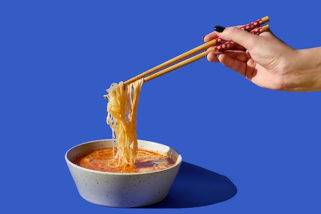 젓가락을 든 여자의 손은 파란색 배경에 대해 그릇에 라면 수프에서 국수를 가져옵니다