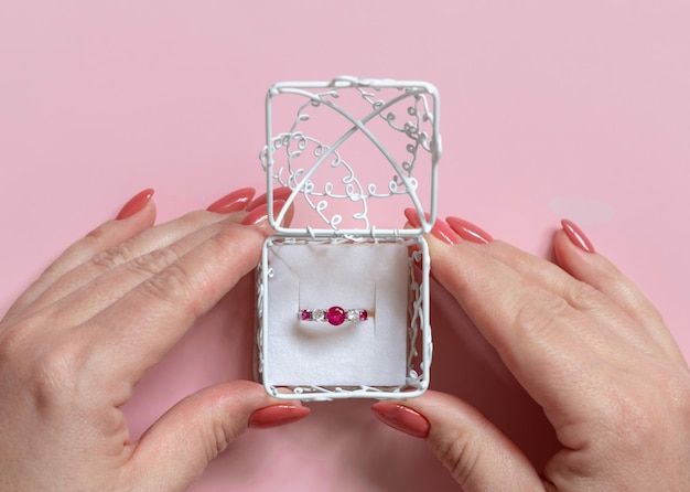 Womans hands opens a ornate gift box with a gemstone engatement ring on pink top view. Marriage proposal concept, valentines day scene