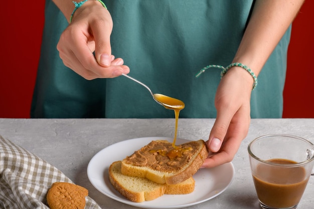 女性の手はスプーンで蜂蜜をピーナッツバターでトーストにめて朝食にサンドイッチを作っています