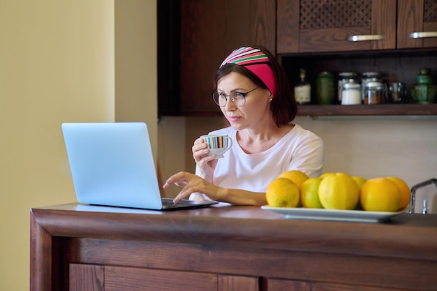 Le donne di 40 anni riposano a casa con una tazza di caffè guardando il laptop