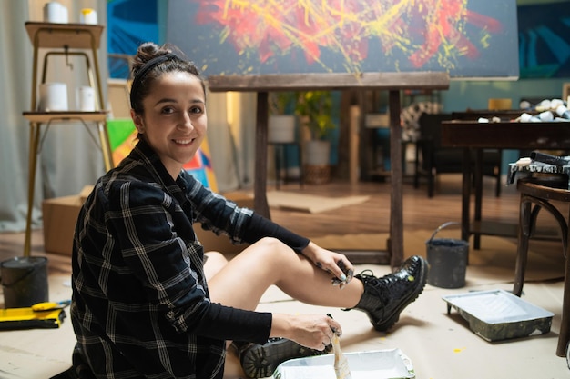 Women in a checkered shirt and clogs is sitting on the floor secured with cardboard among paintings