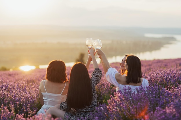 Women clink glasses with white wine in the lavender field at sunset
