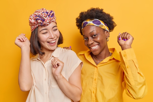 Photo women dance carefree enjoy favorite music keep arms raised up wears shirts smile happily isolated on vivid yellow