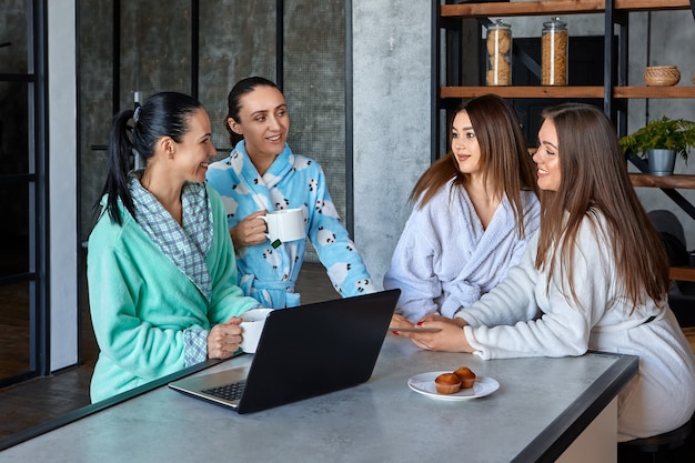 Women in dressing gowns eat breakfast at dining room table and chat while browsing news on laptop computers