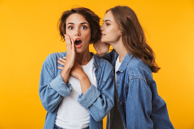 Women friends posing isolated over yellow wall gossiping.