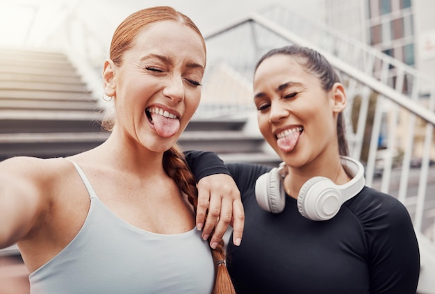 Photo women friends selfie and tongue in city for training comic time or funny face with headphones in profile picture woman workout group exercise photo or social media by stairs in metro with support