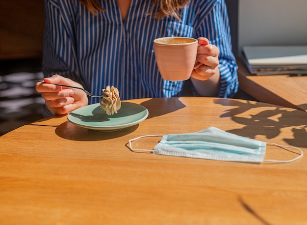 Women hand and coffee cup and sweet dessert and medical face mask on wood desk in coffee house closeup