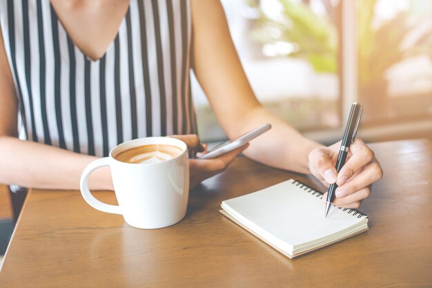 女性は手書きメモ帳にペンで書いて、携帯電話を木製の机で使う。