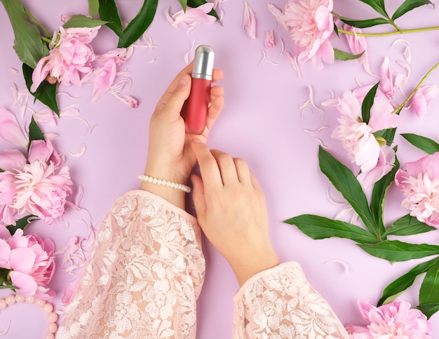 Women hands hold red lipstick, purple background with blooming pink peonies