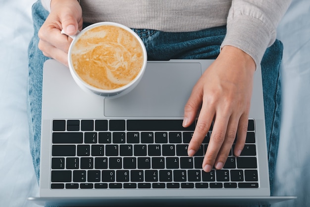 Foto mani delle donne che tengono tazza di caffè latte a casa e controllano il suo laptop