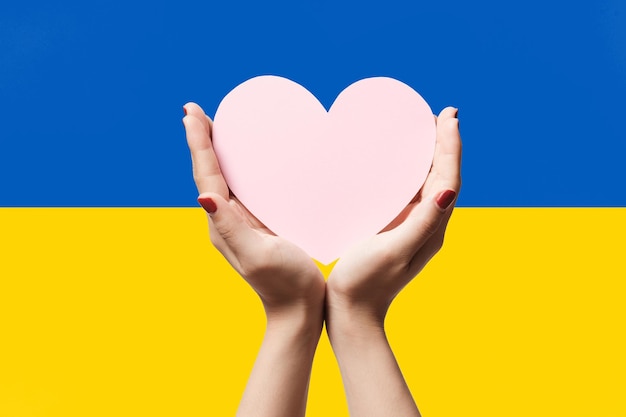 Women hands holding heart shape paper on Ukraine flag color background Stay with ukraine symbol