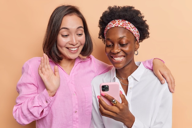 Le donne si divertono a fare videoconferenza tramite smartphone onda ciao ad un amico comunicare a distanza abbracciare e vestirsi casualmente sul muro marrone usare le moderne tecnologie