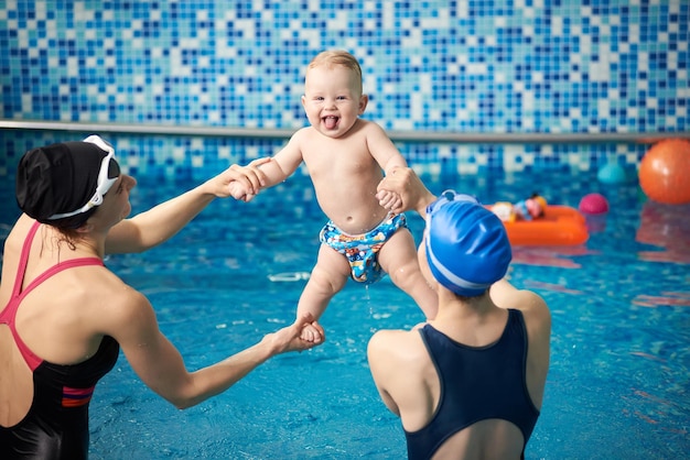 Женщины, держащие малыша, поднимают высоко вверх, растягиваются, тренируются над водой, веселятся в воде, занимаются в бассейне
