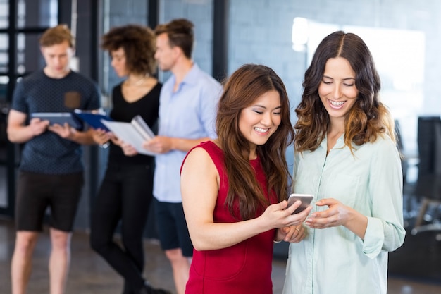 Women looking at smartphone and having discussion while colleagues standing behind in office