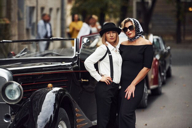 women in old fashioned clothes stands together near retro car.