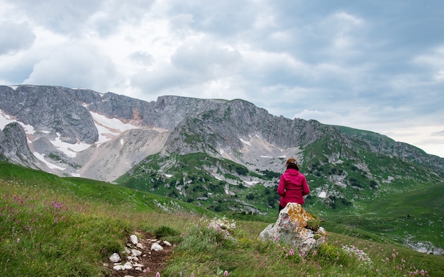 Una donna con una giacca rossa siede su una roccia in una valle di montagna e guarda lontano