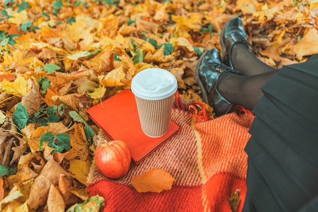 Women resting at blanket on the ground in yellow autumn leaves. cup of coffee red apple and a book. no face