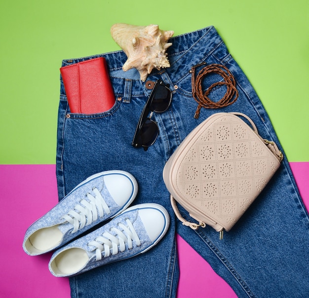 Women's accessories lie on classic jeans with an overstated waist. Sneakers, purse, bag, belt on a pink green background. Top view. The concept of travel.