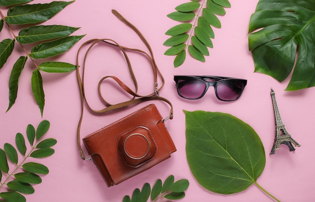 Women's accessories, retro camera, figurine of the Eiffel tower on pink pastel background with green leaves.