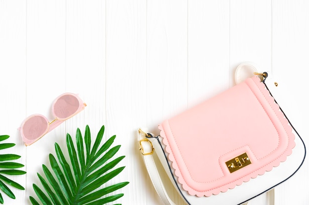 Photo women's accessories - white - pink handbag, trendy rose - colored glasses with mirror glasses, palm leaves on white wooden background