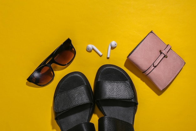 Women's accessories on yellow bright background. Sandals, smart wireless headphone, purses and sunglasses. Top view. Flat lay