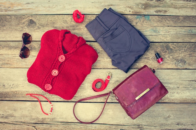  Women's autumn clothing and accessories: red sweater, pants, handbag, beads, sunglasses, nail polish, hair band, belt on wooden background. Toned image. 