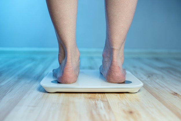 Женские босые ноги стоят на напольных электронных весах, чтобы проверять вес тела и контролировать набор лишних килограммов в синем свете.