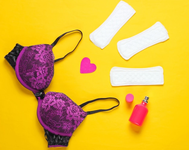 Women's critical days, menstruation. Minimalistic feminine hygiene concept. Beautiful sexy bra, panty liners, perfume bottle, heart on yellow background