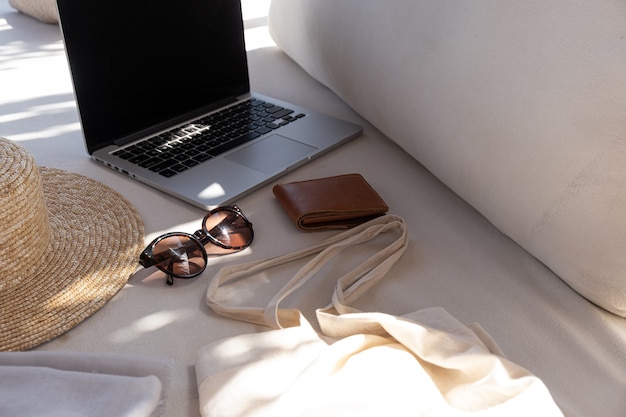 Women's fashion accessories. Stylish female sunglasses, straw hat, shopper bag, laptop on white lounge couch with pillows