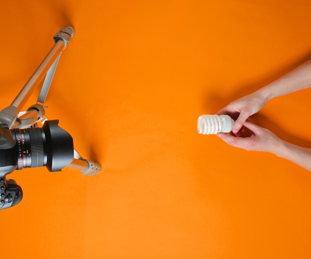 Women's hand review eco spiral light bulb with camera on tripod on orange background. Top view. Minimalism