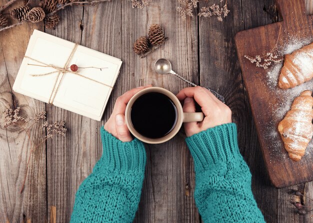 Women's hands in a green knitted sweater holding a ceramic mug with black coffee