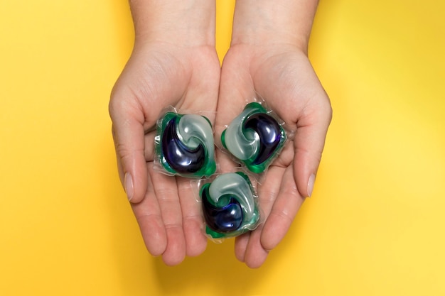 Women's hands hold colored gel capsules for laundry close-up on a yellow background.