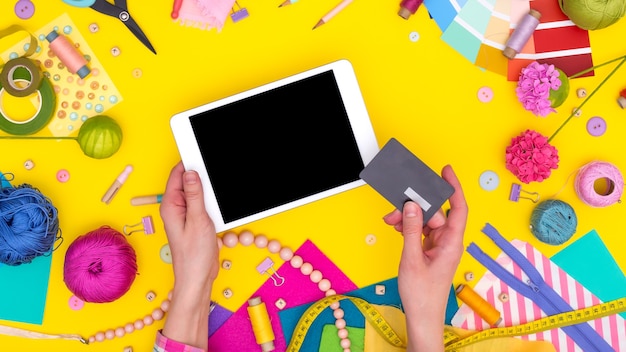 Women's hands hold a credit card and make an online purchase on a tablet. DIY workplace with craft equipment on yellow background. Mock up.