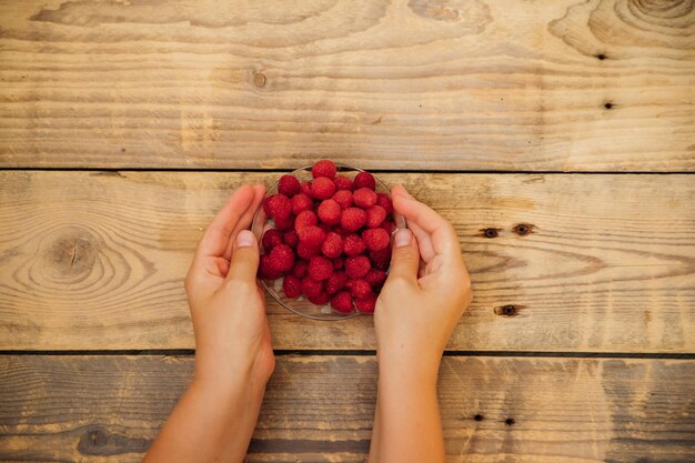 Women's hands hold a small saucer with ripe raspberries a bunch of summer berries on a wooden background Summer background Juicy appetizer or dessert