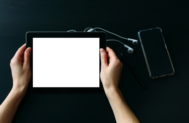 스마트폰과 헤드폰이 있는 검은색 나무 테이블에 빈 흰색 화면이 있는 태블릿 컴퓨터를 들고 있는 여성의 손. 일상 생활에서 디지털 기술의 사용.