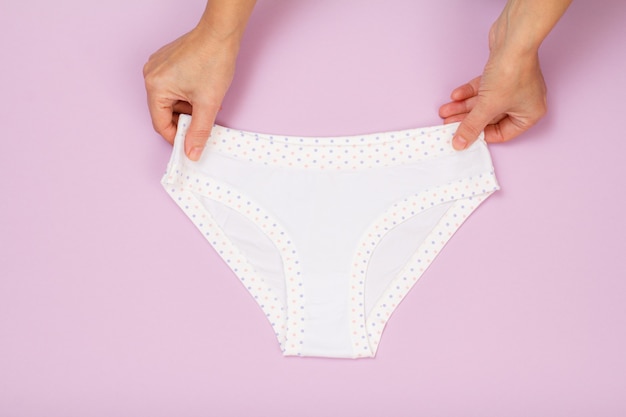 Women's hands holding white cotton panties on lilac background. Woman underwear set. Top view.